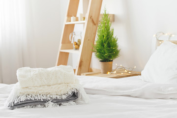 White cozy modern bedroom with holiday decoration. Bed with white bedding set and stack of plaids and sweater on it, wooden rack, Christmas tree in a pot and garland lights. Home christmas decor.