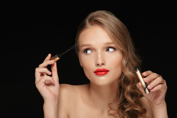 Obraz premium Portrait of young beautiful lady with wavy hair and red lips holding mascara in hand while dreamily looking aside over black background