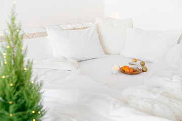 White cozy bedroom with holiday decoration. Bed with white bedding set, wooden plate, cup of tea, tangerines, christmas balls, candle, little Christmas tree and garland lights. Home christmas decor.