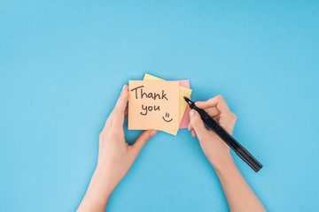 cropped person holding pen above colorful sticky notes with thank you lettering isolated on blue background