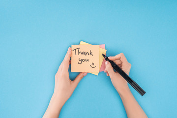 cropped person holding pen above colorful sticky notes with thank you lettering isolated on blue...