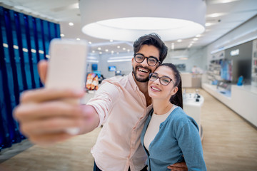 Smiling couple taking selfie while standing in tech store. Contemporary technologies concept.