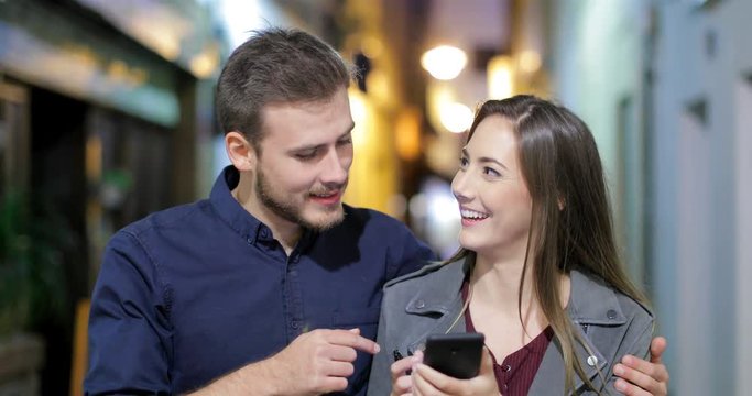 Front view of a happy couple walking using a smart phone in the night in the street of an old town