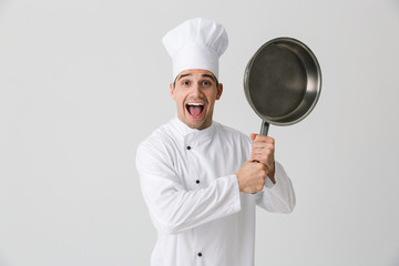 Emotional young man chef indoors isolated over white wall background holding frying pan.