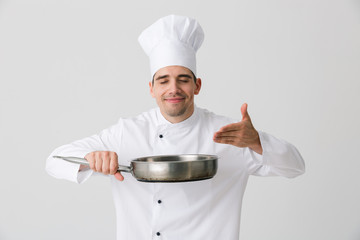 Emotional young man chef indoors isolated over white wall background holding frying pan.