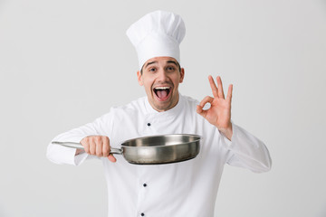 Emotional young man chef indoors isolated over white wall background holding frying pan.