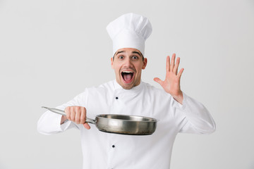 Emotional young man chef indoors isolated over white wall background holding frying pan.