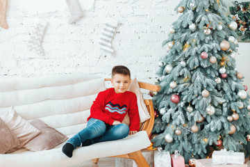 Obraz na płótnie Canvas .Boy is smiling and sitting on the chair near beautiful Christmas tree. New Year Celebration. Decorations. Xmas event