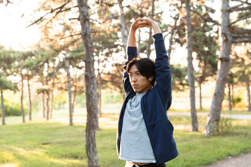 Image of healthy asian guy in casual wear stretching his body, while doing sports in green park