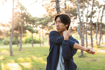 Image of sporty asian guy in casual wear stretching his body, while doing sports in green park