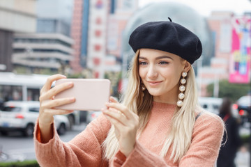 A beautiful young blonde woman dressed in a large warm pink sweater with a black beret and gorgeous earrings. She takes a lot of selfie with her pink smartphone in Chinese city in autumn.