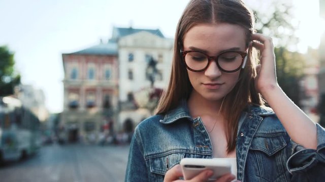 Beautiful young woman with glasses listening to music in headphone use smartphone stand at city on background trem goes sunset look around portrait close up slow motion