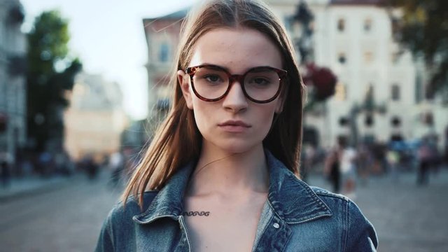Attractive serious young woman stand on the city street look to camera beautiful girl lady jacket lifestyle face summer close up portrait slow motion