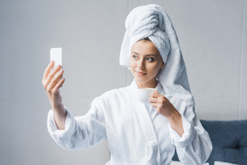 beautiful girl in bathrobe and towel taking selfie with cup of coffee in the morning