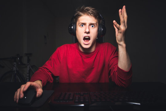 Young gamer is angry with the bad team play in the video game. Portrait of an emotional gamer playing online video games at home on the computer, raising his hands up from surprise.