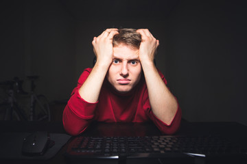 Portrait of a frustrated gamer looking at a computer monitor with a sad faces expression. Young man sits at the table at night at home and plays games on a computer.