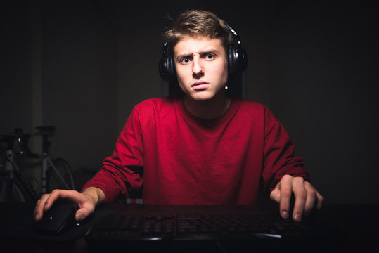 Portrait of a young gamer in headphones sitting at a computer at night and looking at the camera in a concentrated way. Teenager is using the Internet at night on a computer.