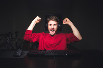 Portrait of an emotional gamer rejoicing with his hands up. Young man sits at night, but uses a...