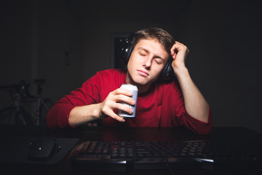 Portrait of a gamer sleeping at the computer at night. Sleepy gamer with a headset sitting at the table with a can of drink in hand and asleep.