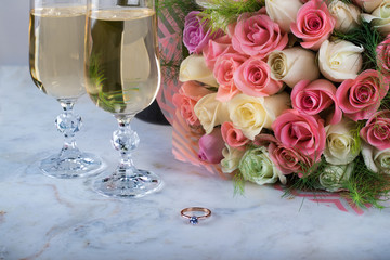 A beautiful bridal bouquet of delicate roses, a ring with a diamond, two glasses of champagne on a marble table. Festive day, wedding. Valentine's Day. A declaration of love, a marriage proposal.