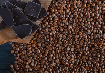 Roasted fragrant beans of black coffee are scattered on a black wooden table, on which black chocolate is lying