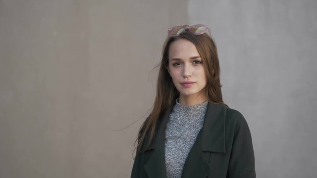 Medium close up of young stylish long hair brunette woman in grey sweater green trench standing on the wind grey city wall. Slow motion hair on wind