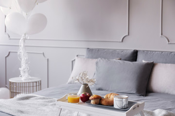 Orange juice, apple, muffins and coffee on wooden trey in stylish grey bedroom interior with bunch of balloons, real photo with copy space