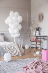 Vertical view of trendy bedroom design with bunch of white balloons, stylish dresser with golden chair and mirror, and pink material on the floor