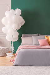Dark green wall with copy space in colorful bedroom with bunch of white balloons next to double bed with grey bedding, pink pillow and blanket