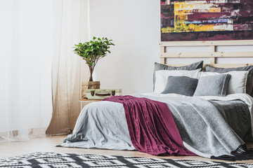 Purple blanket on grey bed and patterned carpet in bedroom interior with plant and painting. Real...