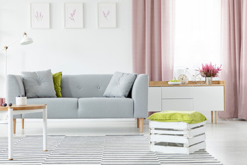 Wooden box with olive green pillow on striped carpet in bright scandinavian living room with grey couch and coffee table, real photo