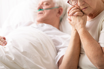 Sad senior woman holding hand of her ill husband lying in hospital bed - obrazy, fototapety, plakaty