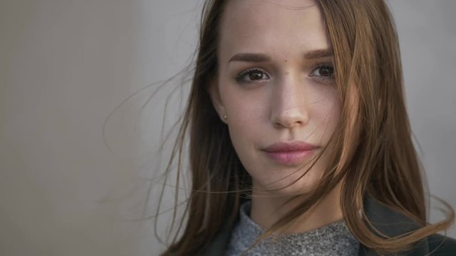 Close up young brunette woman in grey sweater green trench standing on the wind grey city wall fixing hair smiling. Slow motion hair on wind.