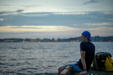 Portrait Asian man sitting on the port and looking out to the sea with the twilight sky of sunset and bokeh lights of the city in background