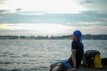 Portrait Asian man sitting on the port and looking out to the sea with the twilight sky of sunset and bokeh lights of the city in background