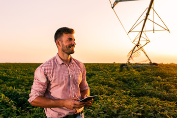 Young farmer holding tablet in his hands and adjusts irrigation system on soybean field at sunset. © Zoran Zeremski