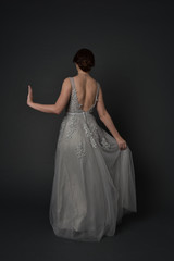 full length portrait of brunette girl wearing beautiful long gown, standing pose with background to the camera on grey studio background.