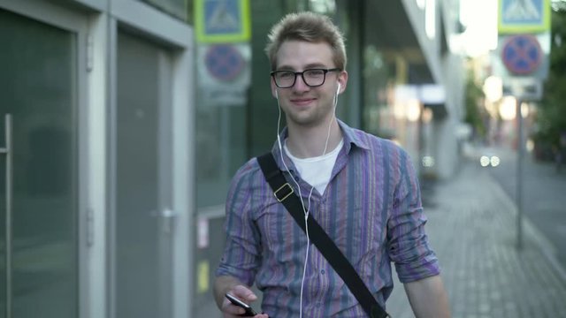 Cheerful young man wearing striped shirt and glasses walking in the street and listening to music in his headphones. Tracking real time medium shot