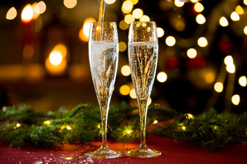 Background of two glasses of champagne, spruce branches on red table cloth. Living room decorated with lights and candles and Christmas tree