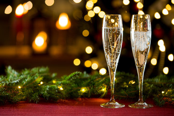 Background of two glasses of champagne, spruce branches on red table cloth. Living room decorated with lights and candles and Christmas tree
