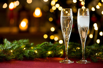 Background of two glasses of champagne, spruce branches on red table cloth. Living room decorated with lights and candles and Christmas tree