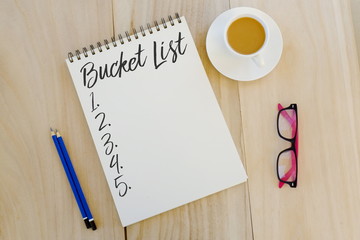 Top view of pencil,glasses,a cup of coffee and notebook written with Bucket List on wooden background.