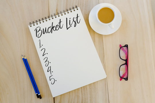 Top view of pencil,glasses,a cup of coffee and notebook written with Bucket List on wooden background.