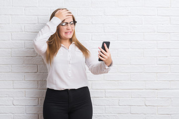 Young adult business woman over white brick wall sending message using smartphone stressed with hand on head, shocked with shame and surprise face, angry and frustrated. Fear and upset for mistake.