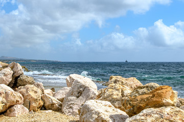 Seascape with huge rocks in the foreground, surf waves, ship silhouette on the horizon and blue sky with clouds.