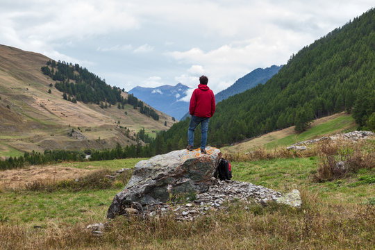 Man wearing a red technical jacket standing on a rock, looking at the landscape on italian Alps on a cloudy day in Valle Varaita, Piemonte (Piedmont), Northern Italy,