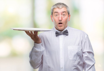 Handsome senior waiter man holding silver tray over isolated background scared in shock with a...