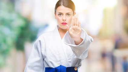 Young beautiful woman wearing karate kimono uniform over isolated background doing stop sing with palm of the hand. Warning expression with negative and serious gesture on the face.