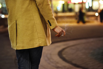 Elegant fashion man wearing modern coat, standing on street at night and smoke a cigarette. Man...