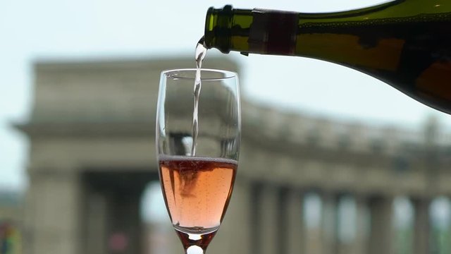 Pouring pink champagne or sparkling wine to glass from bottle. European city on background. Slowmotion